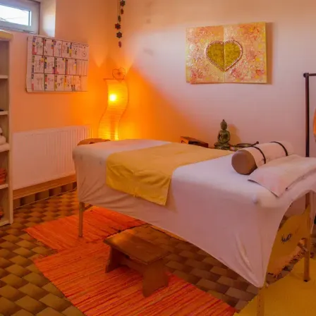 Ein Massageraum mit einem Bett, einer Lampe und einem Gemälde an der Wand. Ein Regal und eine Heizung sind sichtbar. Der Boden ist gefliest, und der Raum ist warm.