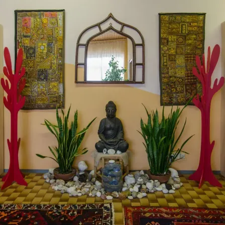 Ein Zimmer mit einer Buddha-Statue, zwei Pflanzen, einem Spiegel und dekorativen Wandbehängen. Es gibt zwei rote Bäume auf den Seiten und einen Teppich auf dem Boden.