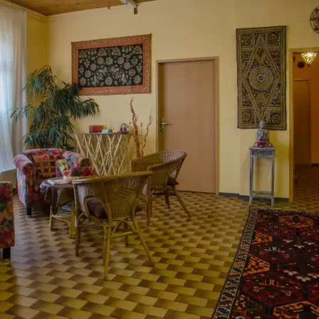 Ein gemütliches Wohnzimmer mit gelben Wänden, einer Holztür und einem gefliesten Boden. Es gibt Stühle und einen Tisch mit dekorativen Gegenständen. Zwei Teppiche hängen an der Wand.
