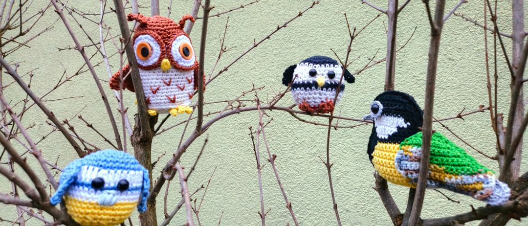 Vier gehäkelte Vögel, darunter eine Eule und drei andere, sitzen auf einem kahlen Ast einer grünen Wand.
