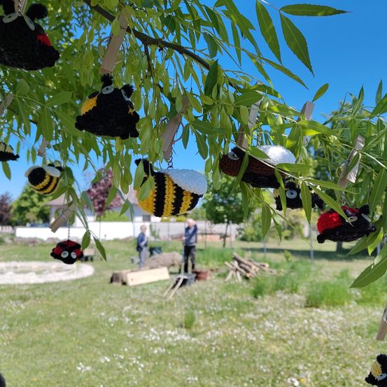 Handmade knitted bee keychains hang from a tree. Two people work in the background. The sky is clear, and there is a house in the distance.