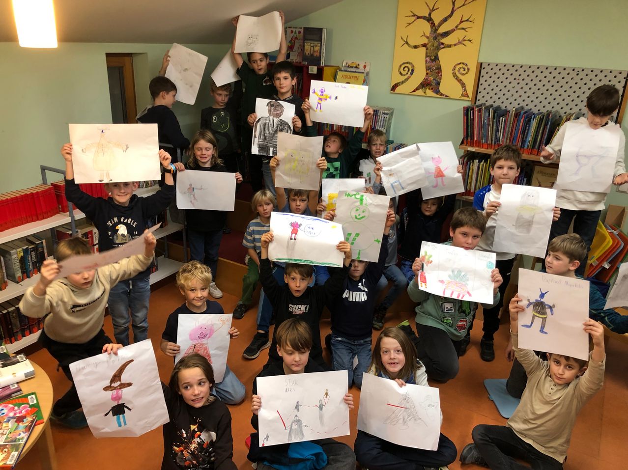 Ein Klassenzimmer voller Kinder, die ihre Zeichnungen auf weißen Blättern hochhalten, mit einigen Büchern und einem Baumbild im Hintergrund.