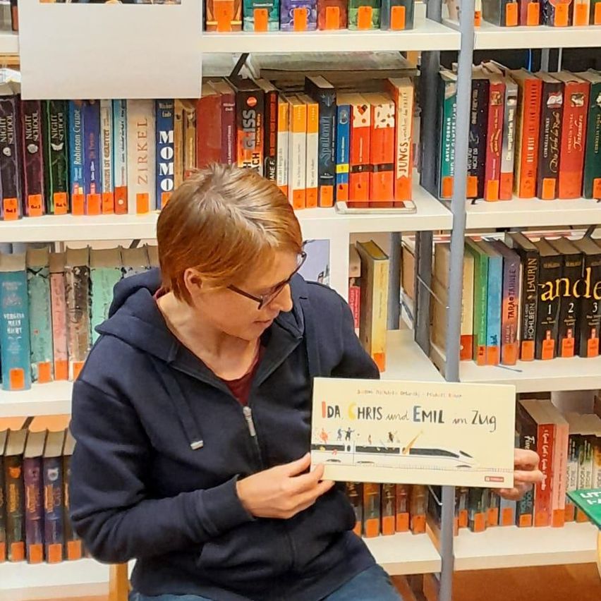 Eine Person mit Brille und einer dunkelblauen Kapuzenjacke hält ein Buch mit dem Titel 'IDA CHRIS und EMIL an Zug' in einer Bibliothek.