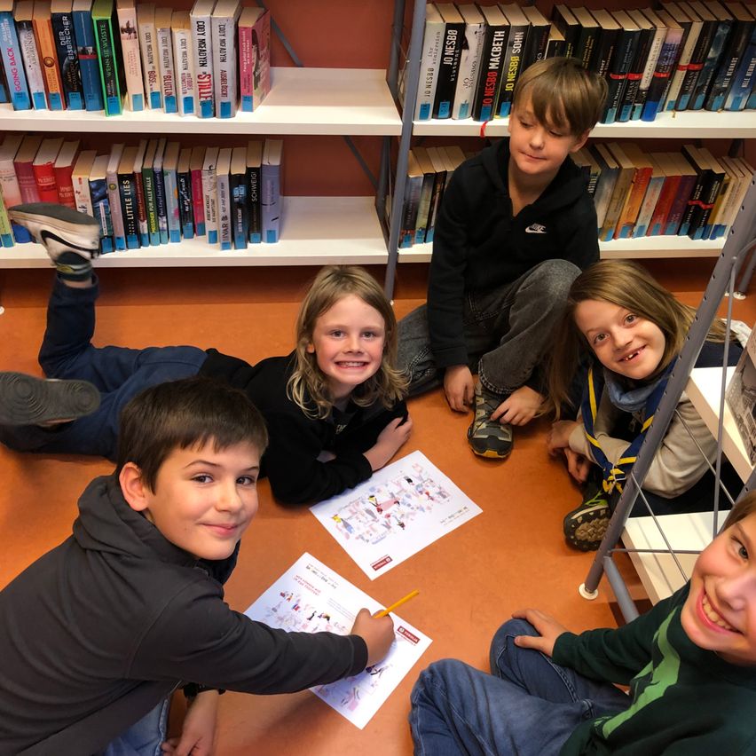 Eine Gruppe von Kindern sitzt auf dem Boden in einer Bibliothek, lächelt und schaut auf ein Stück Papier. Im Hintergrund befinden sich mit Büchern gefüllte Bücherregale.