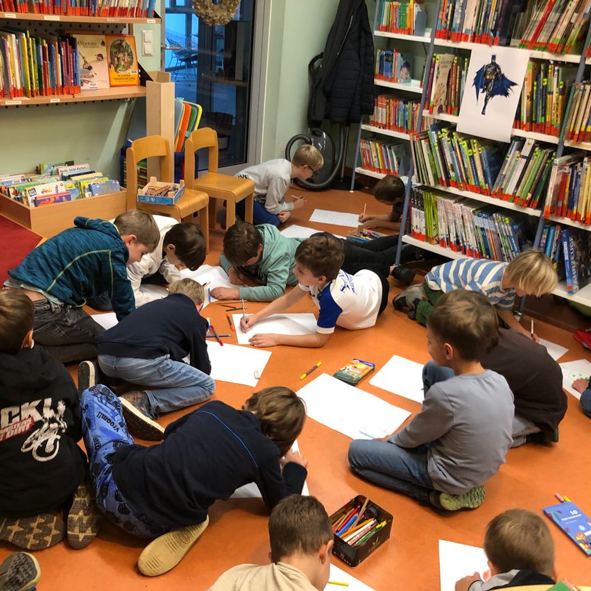 Eine Gruppe von Kindern sitzt auf dem Boden in einer Bibliothek und zeichnet auf großen Papierblättern. Einige benutzen Stifte, andere Kreiden. Der Raum ist mit Büchern an den Regalen und einem Batman-Poster gefüllt.