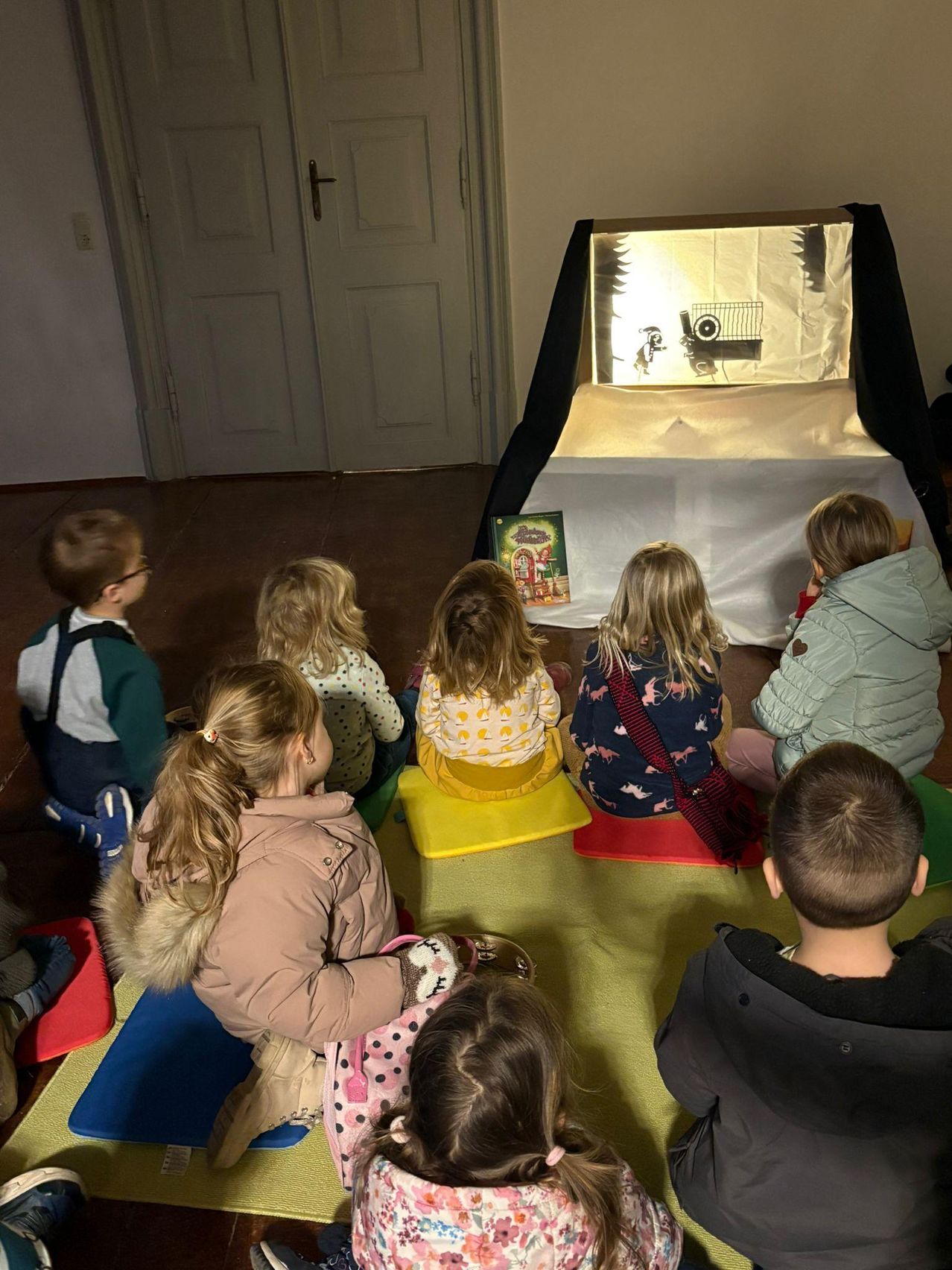 Eine Gruppe von Kindern schaut sich eine Puppentheateraufführung auf einer Bühne in einem Raum an. Sie sitzen auf Kissen auf dem Boden.