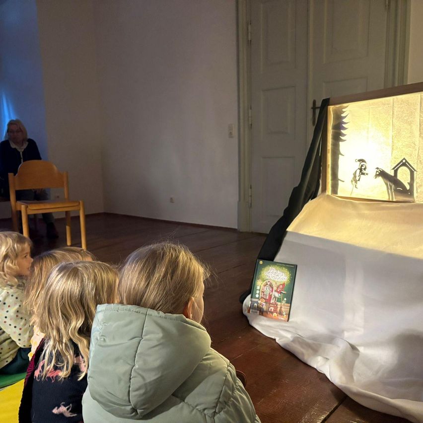 Kinder schauen sich eine Schattenspielvorstellung in einem Raum mit einem auf einem weißen Tuch ausgestellten Buch an. Eine Frau sitzt in der Nähe auf einem Stuhl.