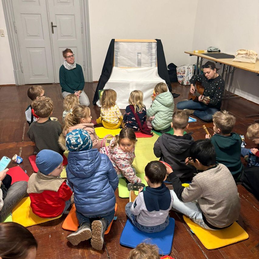 In einem Klassenzimmer sitzt eine Gruppe von Kindern im Kreis auf dem Boden und lauscht einer Frau, die Gitarre spielt. Die Frau kniet vor ihnen, und die Kinder sind aufmerksam und beobachten sie.