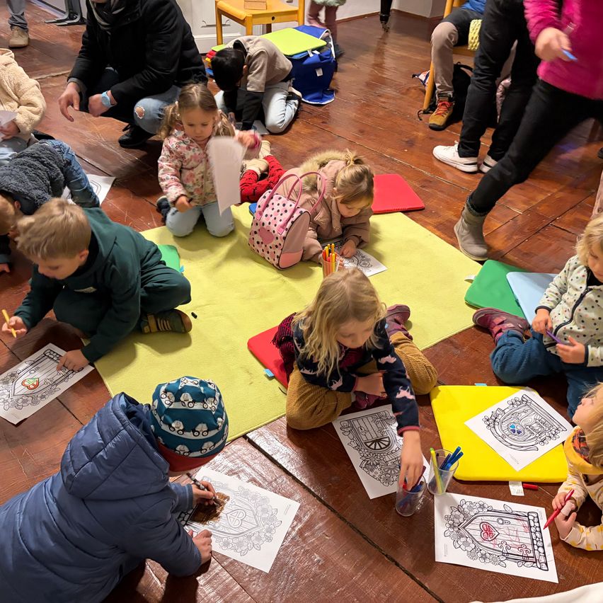 Eine Gruppe von Kindern und Erwachsenen malt auf einem Holzboden. Sie sitzen auf dem Boden mit gelben Matten und halten Malstifte. Einige tragen Winterkleidung.