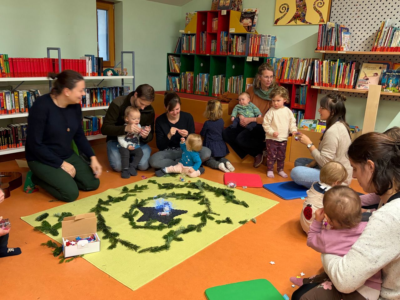 In einer Bibliothek sitzen eine Gruppe Erwachsener und Kinder auf dem Boden um einen kreisförmigen Teppich mit Grünpflanzen. Einige halten Babys und Spielzeug. Regale mit Büchern umgeben sie.
