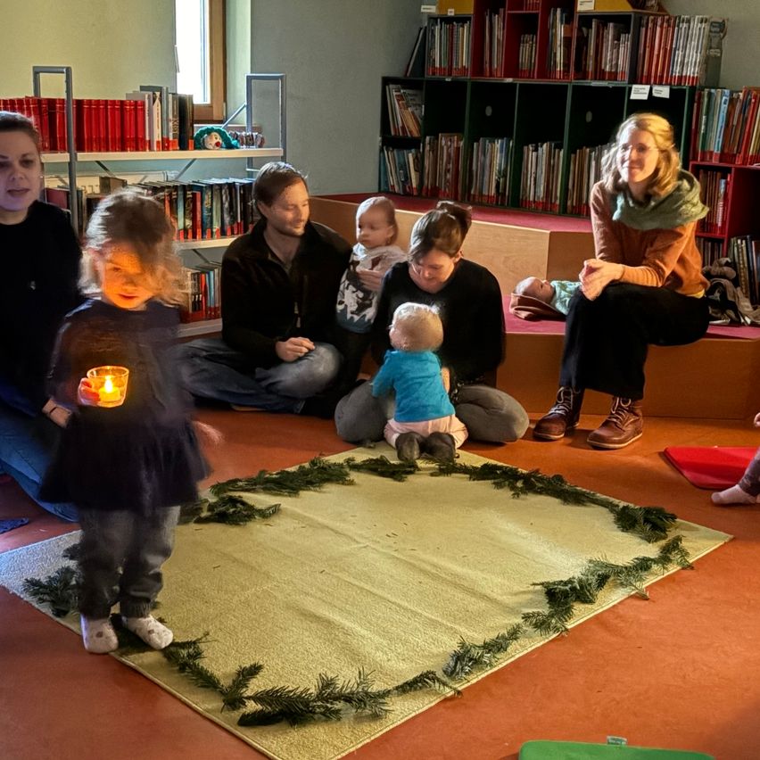 Eine Gruppe von Erwachsenen und Kindern sitzt im Kreis auf einem Teppich, mit einem kleinen Kind, das eine Kerze in der Mitte hält. Sie sind in einem Raum mit Regalen voller Bücher.
