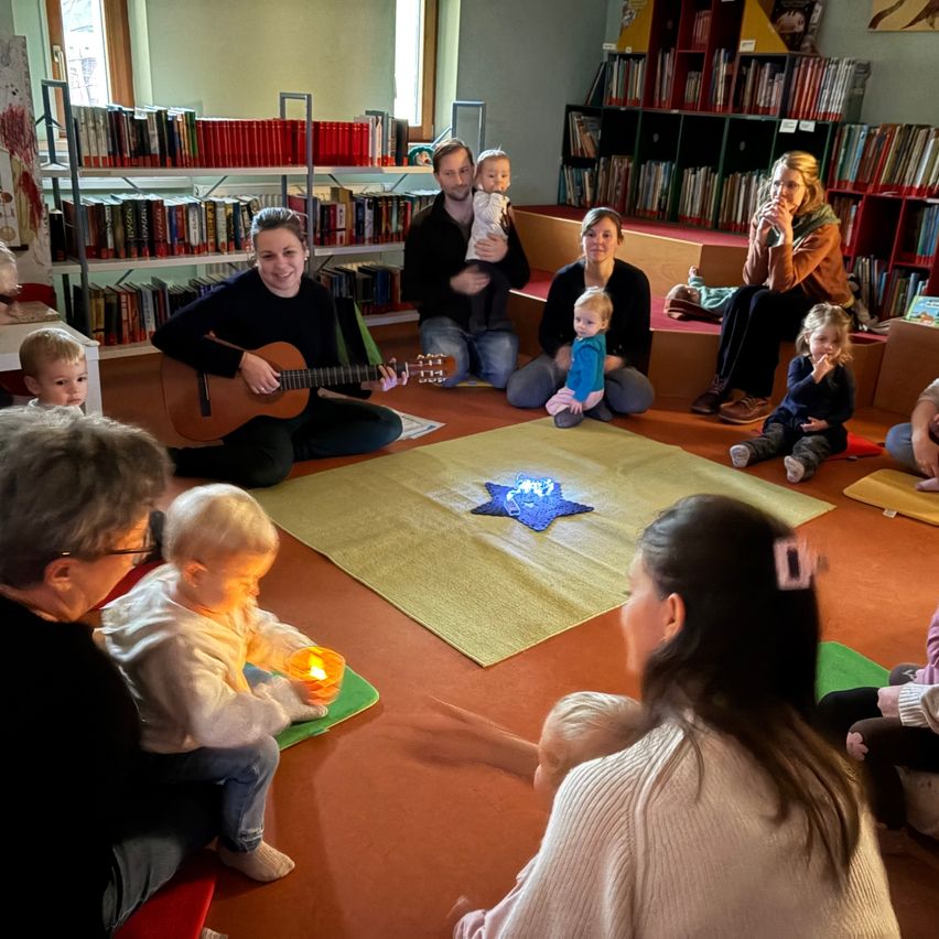 In einem Raum sitzen mehrere Erwachsene und Kinder in einem Kreis um einen Stern. Eine Frau spielt Gitarre. Die Kinder spielen mit einer Kerze. Hinter ihnen befinden sich Bücherregale mit Büchern.