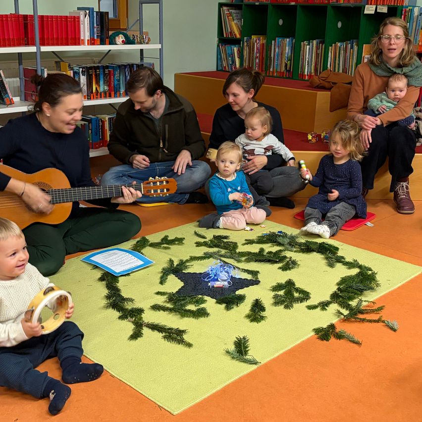Eine Gruppe von Erwachsenen und Kindern sitzt in einer Bibliothek auf dem Boden, spielt Instrumente und singt. Ein Teppich mit einem Kranzdesign liegt auf dem Boden.