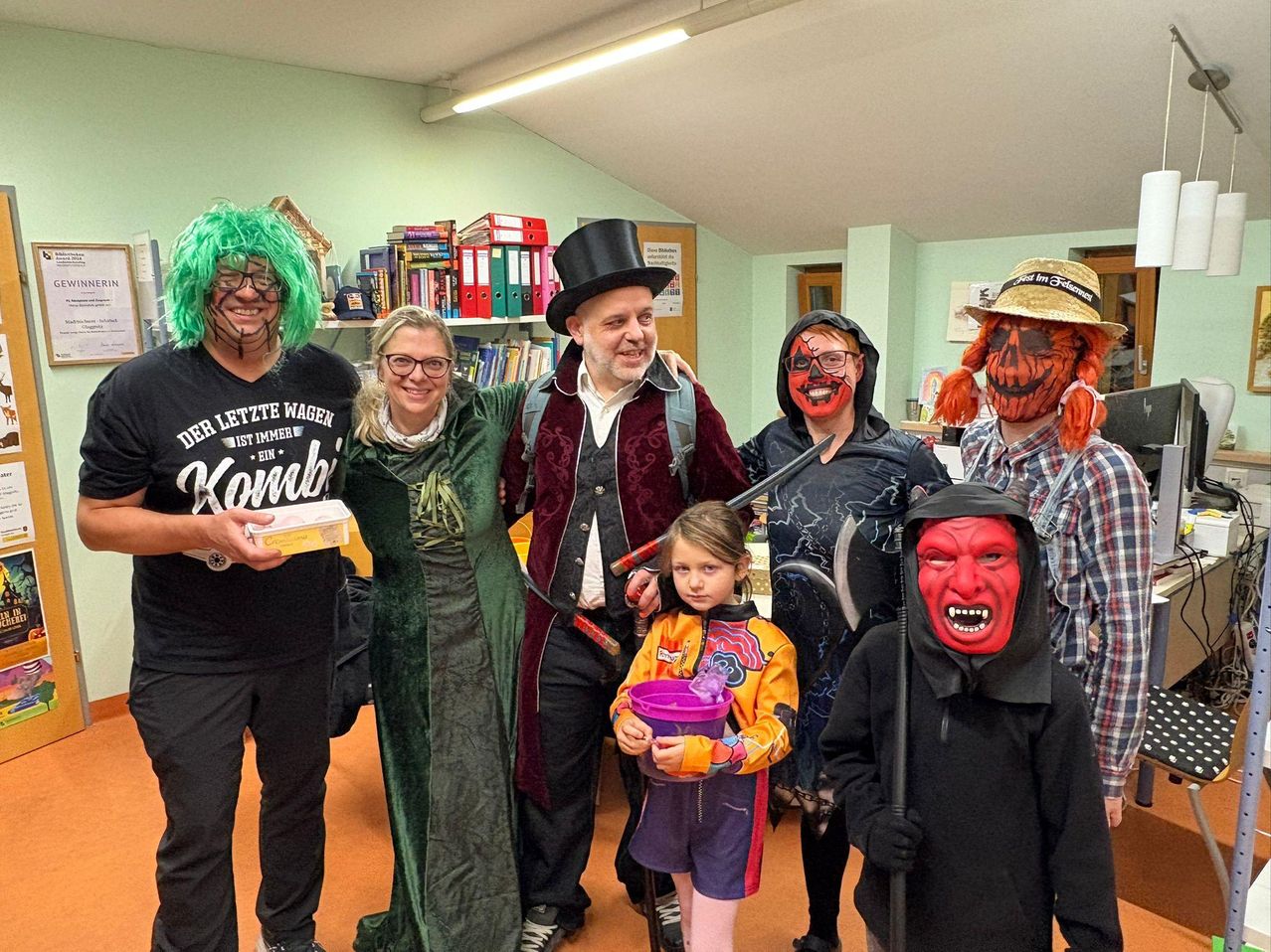 Eine Gruppe von Menschen in Kostümen steht in einem Raum. Ein Mann mit Topfhut, eine Frau in einem grünen Kleid und ein junges Mädchen mit Eimer.