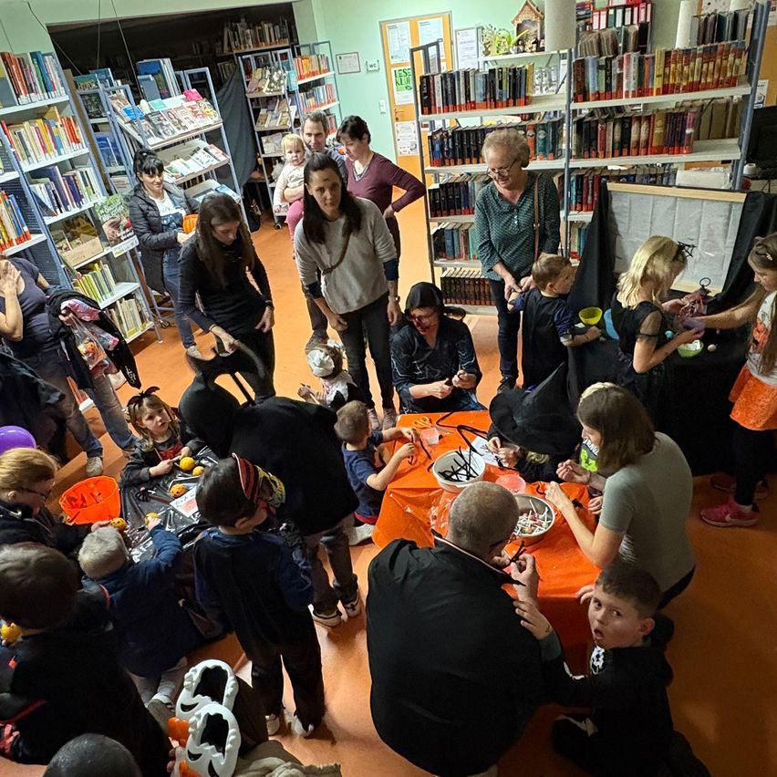 Eine Gruppe von Kindern und Erwachsenen trifft sich in einer Bibliothek für eine Halloween-Veranstaltung, trägt Kostüme und beteiligt sich an Aktivitäten rund um einen orangefarbenen Tisch.
