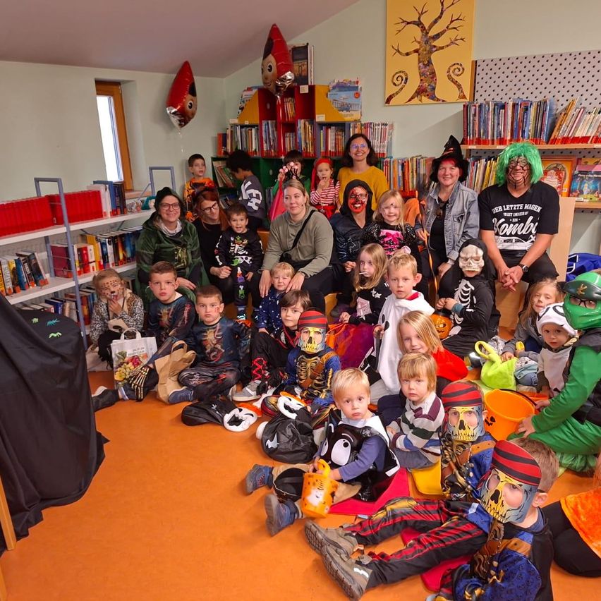 Eine Gruppe von Kindern in Halloween-Kostümen sitzt auf dem Boden in einem Raum, umgeben von Erwachsenen, mit Regalen voller Bücher im Hintergrund.