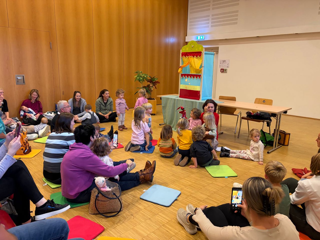 A group of adults and children are seated on the floor in a room, watching a puppet show. The children are seated on colorful mats, and the adults are sitting on the floor or chairs. A woman is holding a cell phone and a stuffed toy. The room has wooden floors and walls.