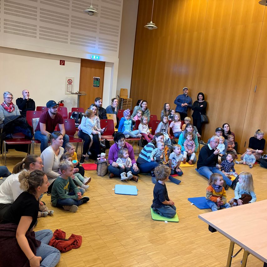 A group of people, including adults and children, are gathered in a room. Some are sitting on the floor, while others are standing. Some of the children are holding toys. The room has wooden walls and a wooden floor.