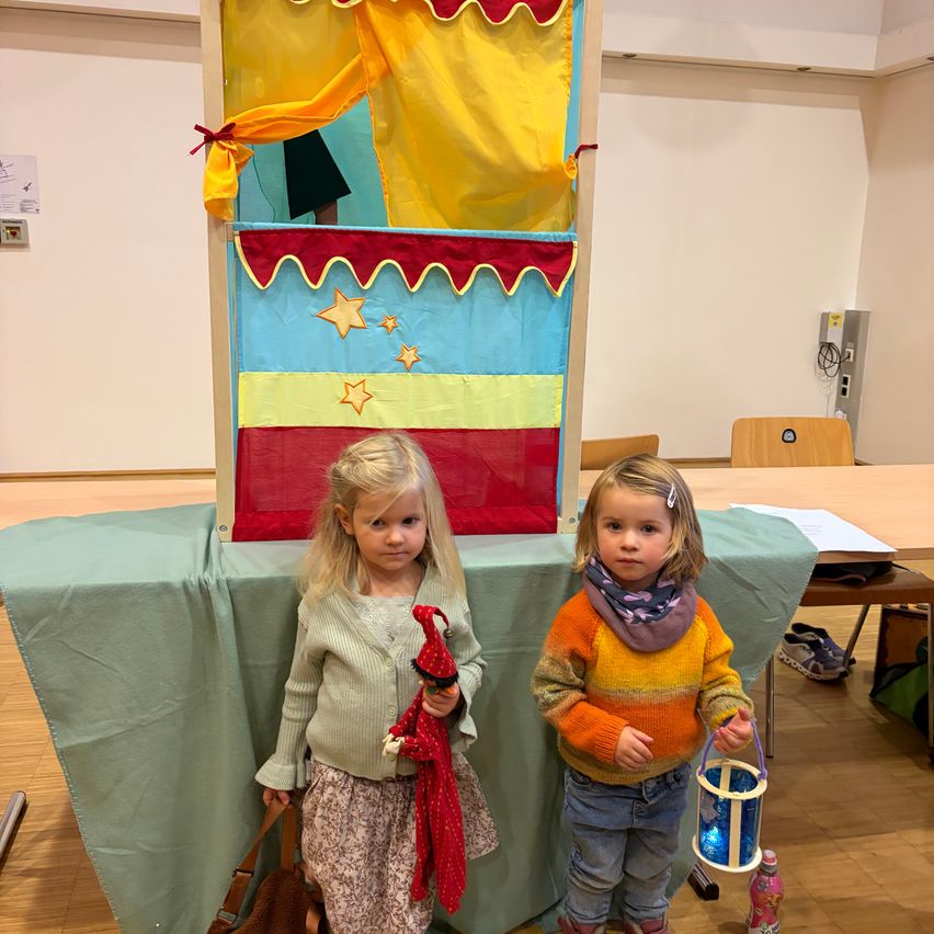 Two young girls stand in front of a colorful puppet theater on a table. One holds a red puppet, the other a lantern. The theater has a yellow curtain and stars. A green tablecloth covers the table.