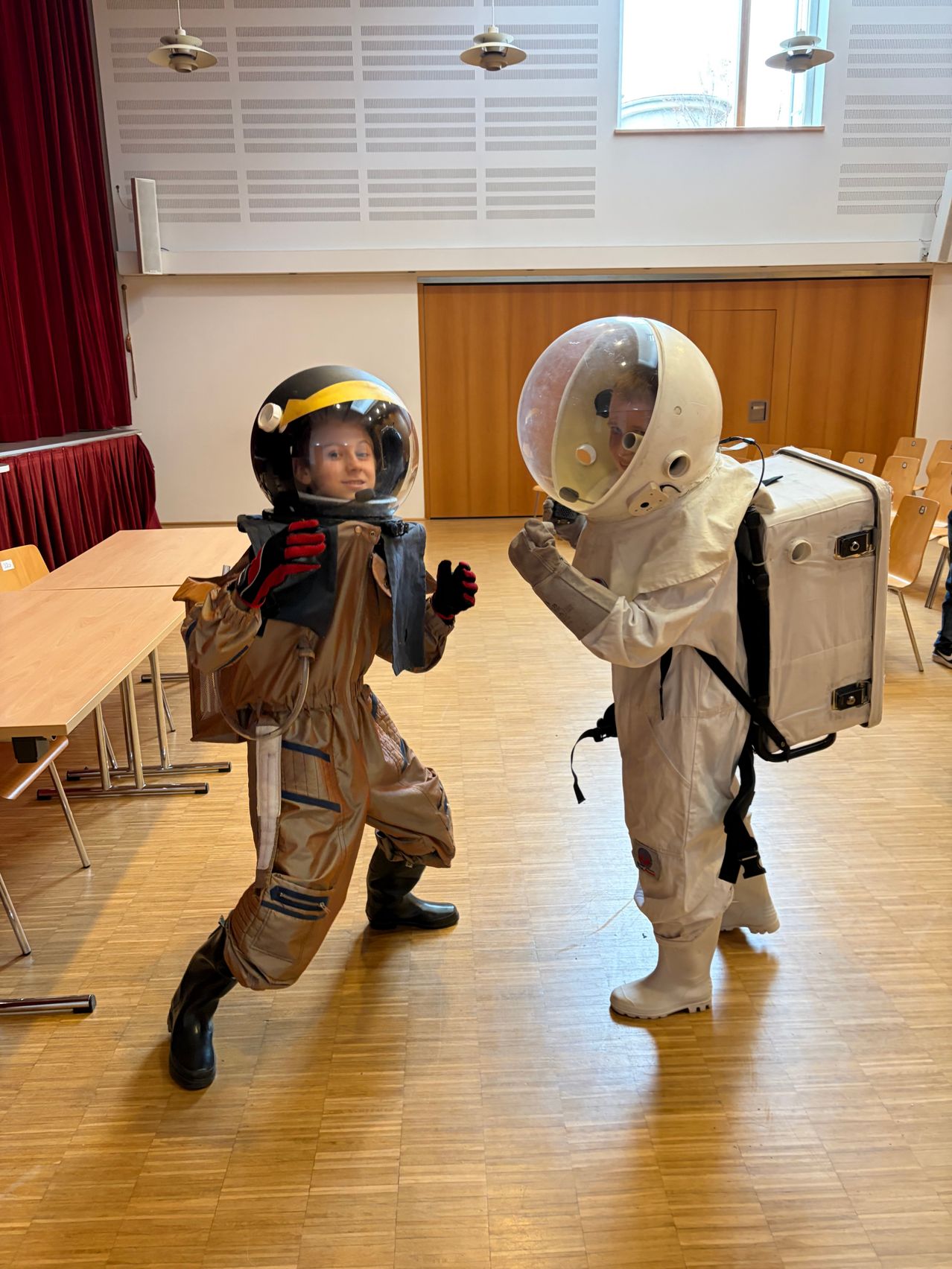 Two kids dressed as astronauts face each other in a room with wooden floors and chairs.