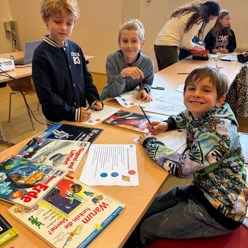 Three boys sit at a table, engrossed in coloring a worksheet. One boy holds a pen and the other holds a marker. Behind them, a woman stands at another table.