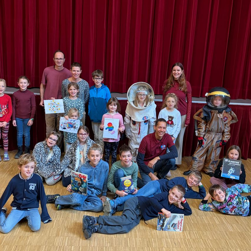 A group of children and adults posing for a photo with some dressed in astronaut costumes. Two individuals in astronaut suits stand at the sides.