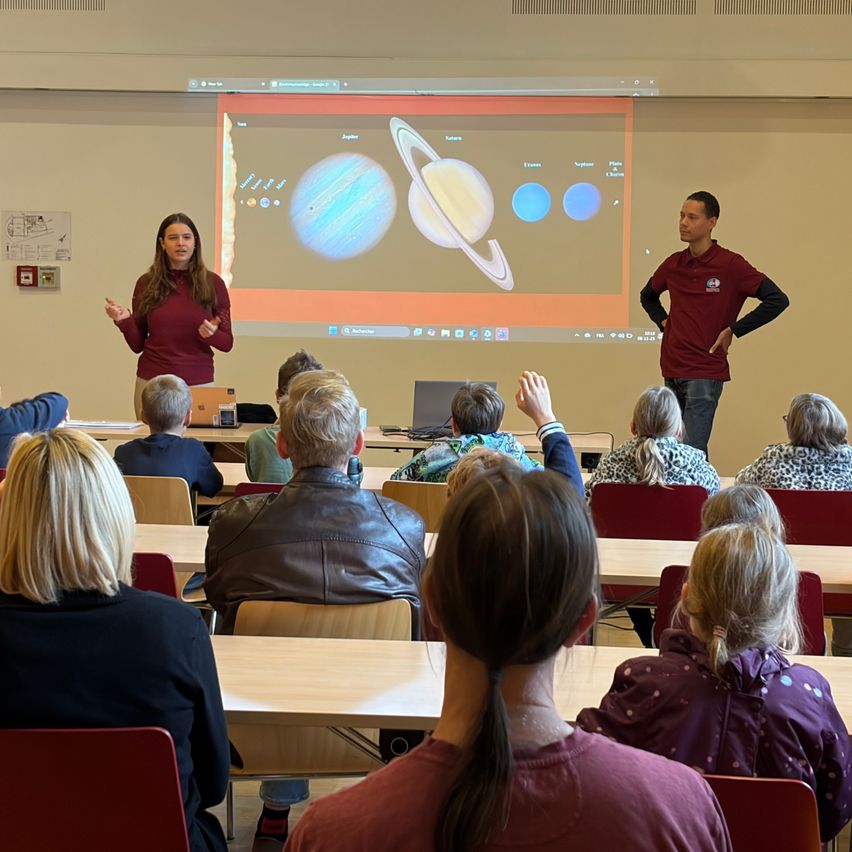 A classroom setting with two presenters standing in front of a projector screen showing images of planets. Students sit attentively in chairs, some raising their hands.