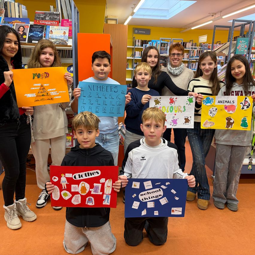 Kinder in einer Bibliothek posieren mit pädagogischen Postern über Essen, Zahlen, Farben, Tiere und Schulsachen.