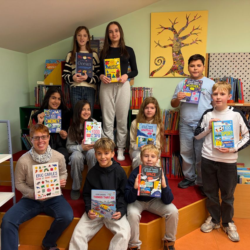 Eine Gruppe von Kindern und Erwachsenen posiert mit Büchern in einer Bibliothek. Die Kinder halten bunte Bücher in den Händen und die Erwachsenen stehen hinter ihnen. Die Bibliothek ist gefüllt mit Bücherregalen und einem farbenfrohen Gemälde an der Wand.