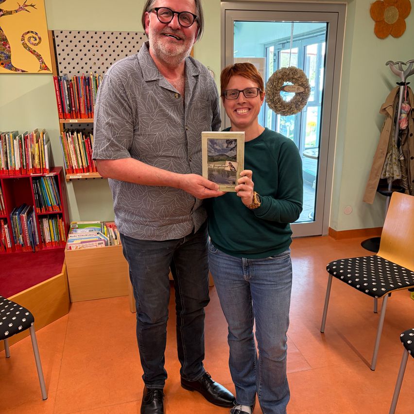 A man and a woman stand in a room, smiling. The man holds a framed picture. Behind them, a bookshelf and a door with a wreath are visible.