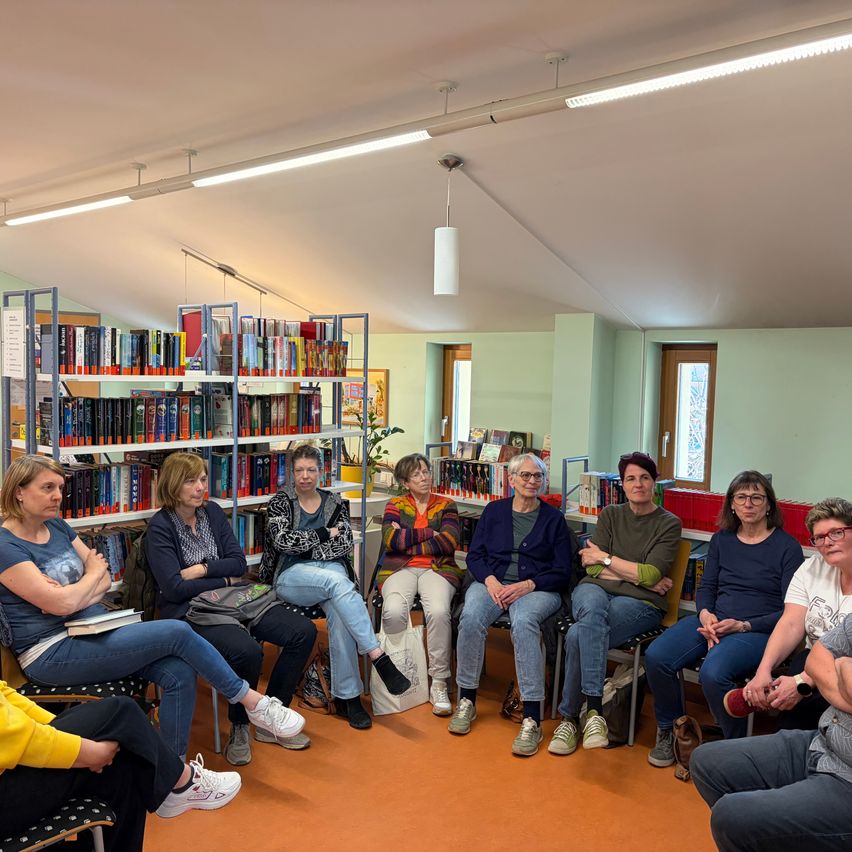 A group of women sit on chairs in a library with bookshelves in the background. Some are wearing glasses, and there is a plant and a lamp in the room.
