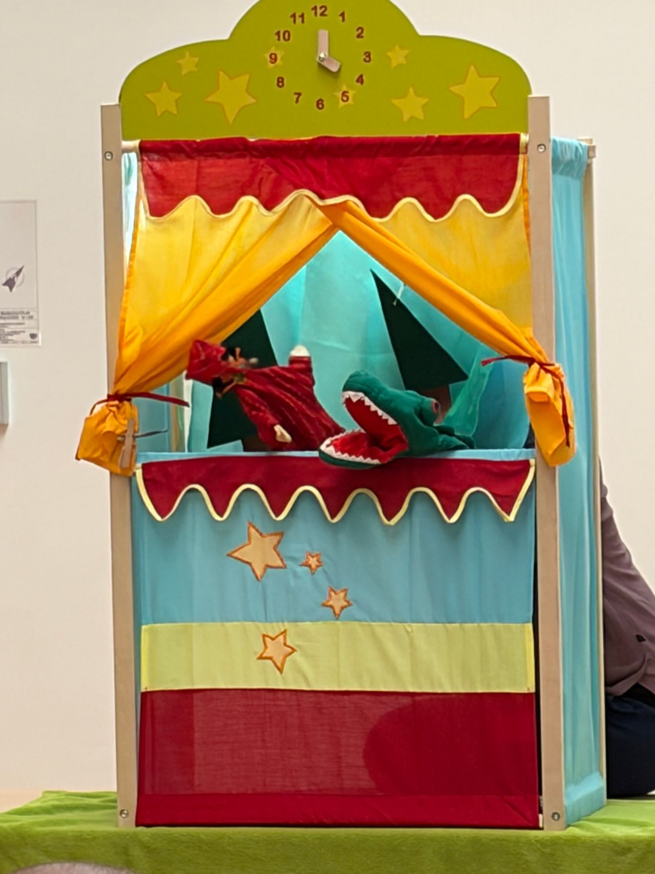 A colorful puppet theater with a red and yellow curtain. Inside, a red puppet and a green crocodile puppet are staged, with a blue backdrop featuring stars.