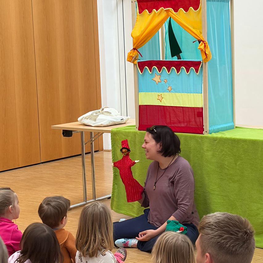 A woman is sitting on the floor with a puppet on a stage with a colorful curtain, entertaining children.