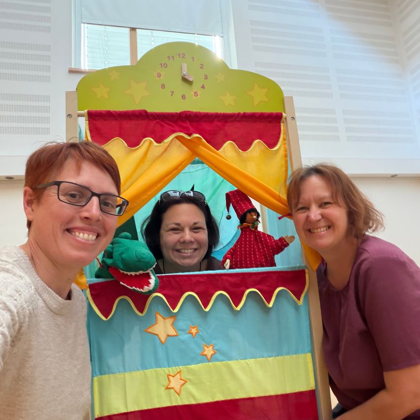 Three women are posing for a photo inside a colorful puppet theater, with a puppet and a green crocodile. A clock is visible on the top.