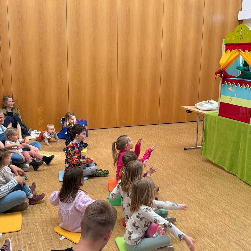 A group of children and adults are sitting on cushions in a room watching a puppet show on a stage.