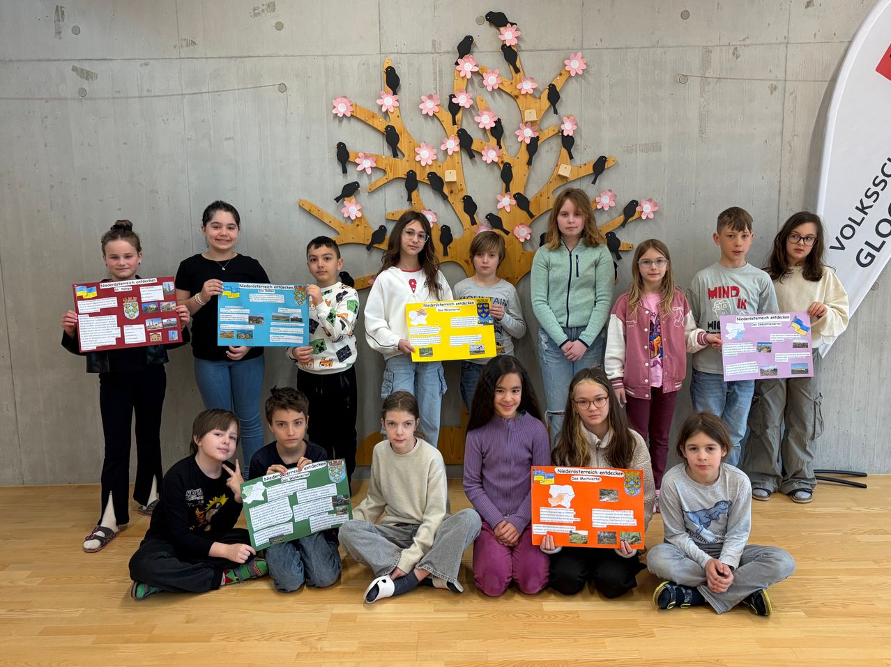 A group of children and young people are posing for a photo while holding colorful posters. They are smiling and standing in a room with a tree backdrop.