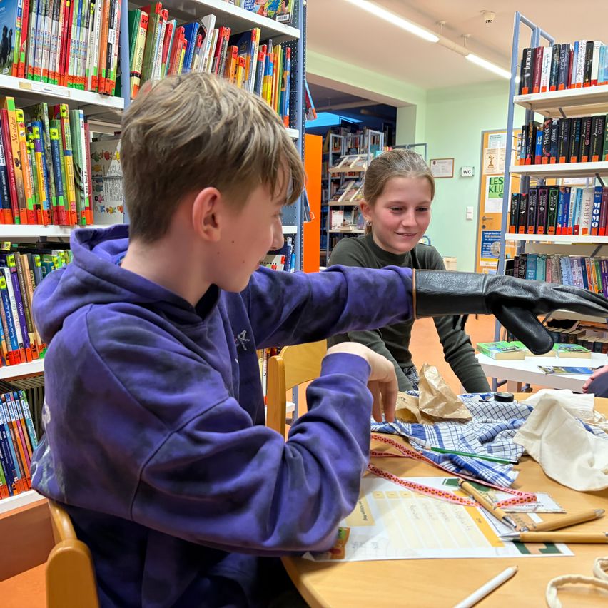 Zwei Kinder arbeiten in einer Bibliothek an einem Bastelprojekt, umgeben von Büchern und Zubehör.