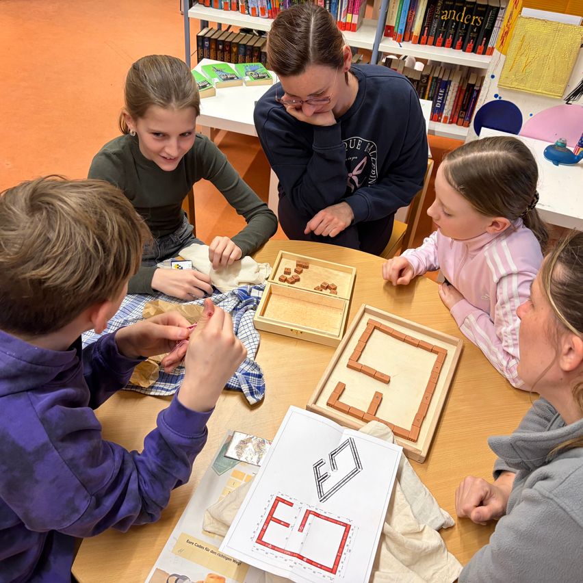 Mehrere Kinder sitzen um einen Tisch mit Puzzleteilen. Eine Frau steht hinter ihnen und sieht sie an. Der Raum ist eine Bibliothek mit Regalen voller Bücher.