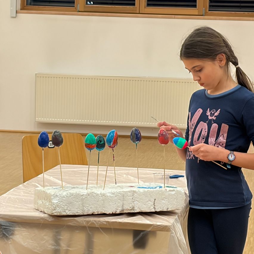 Ein Mädchen steht vor einem Tisch mit einem Schaumstoffblock und farbigen Eiern und malt sie. Sie hält einen Pinsel in der rechten Hand.
