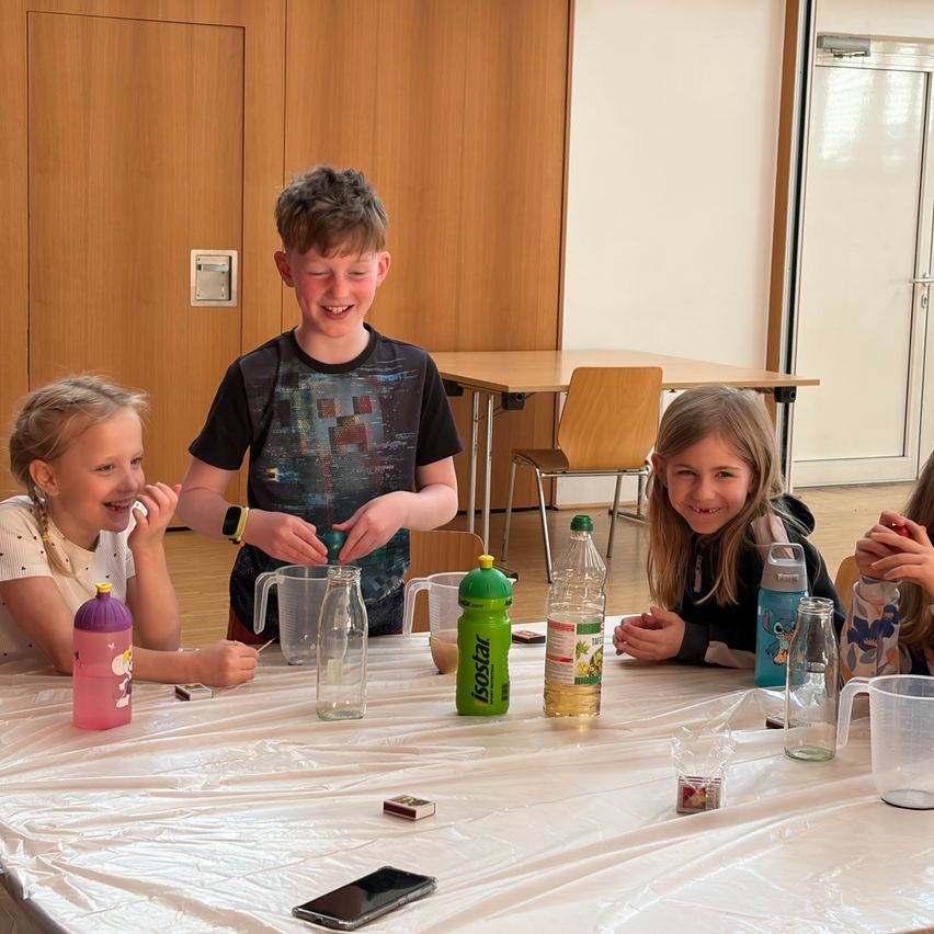 Eine Gruppe von Kindern sitzt lächelnd um einen Tisch. Ein Junge fügt etwas zu einem Krug hinzu. Flaschen, ein Telefon und andere Gegenstände sind auf dem Tisch. Der Raum hat Holzwände und Türen.