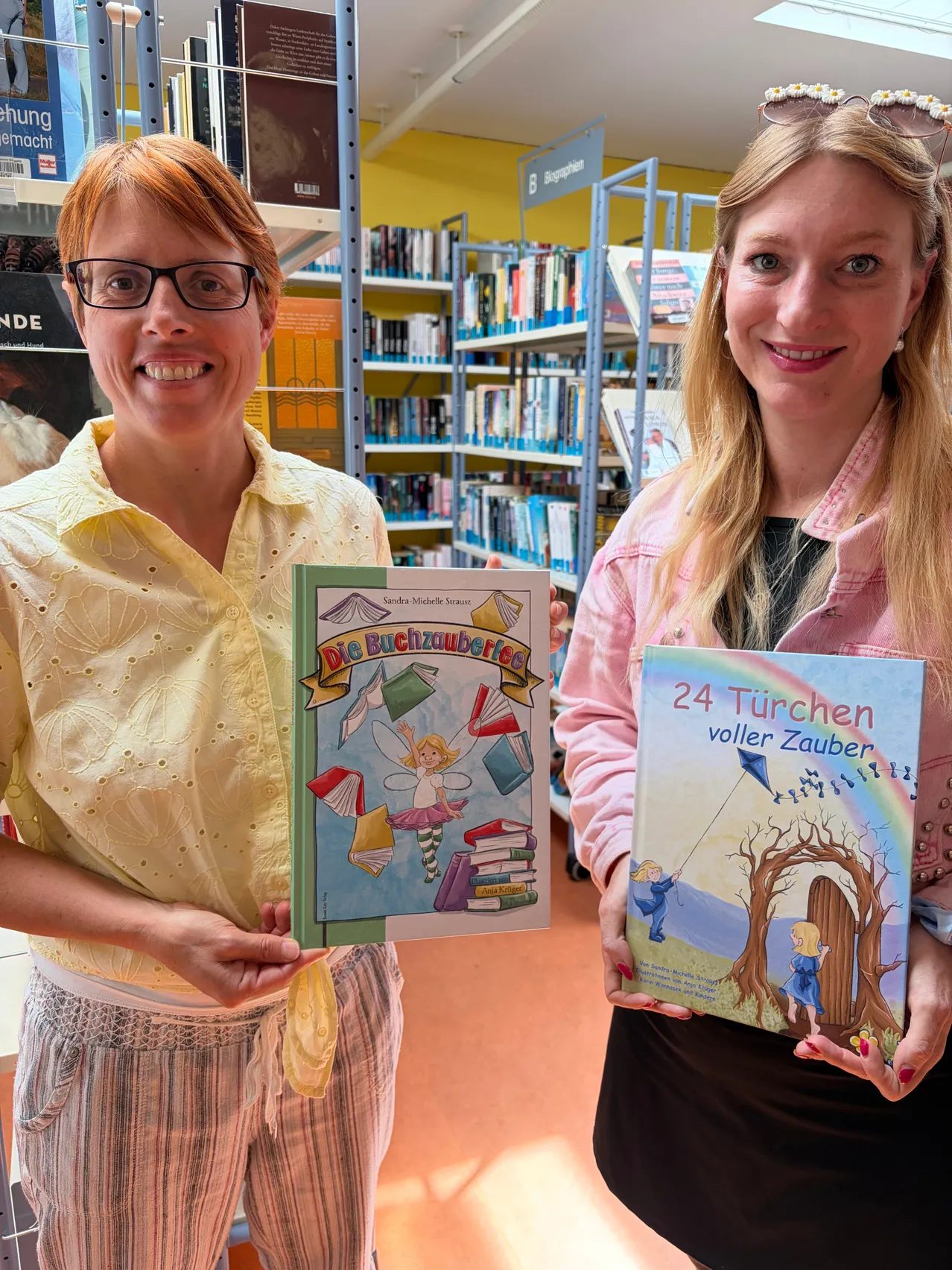 Zwei Frauen in einer Bibliothek, eine hält ein Buch mit dem Titel 'Die Buchzauberei' und die andere hält ein Buch mit dem Titel '24 Turchen voller Zauber'. Sie lächeln und posieren für ein Foto.