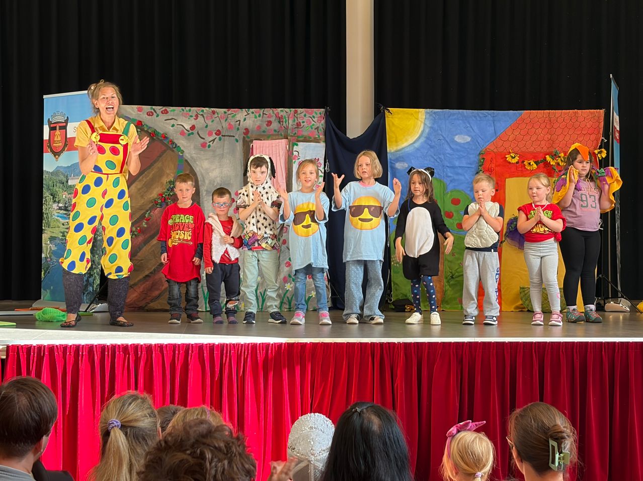 Eine Gruppe von Kindern in verschiedenen Kostümen steht auf der Bühne mit einer Frau in einem Clownskostüm. Sie führen vor einem Publikum auf.