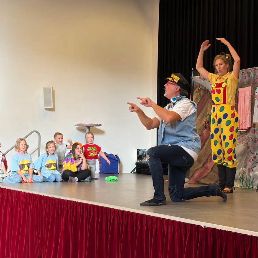 Ein Clown und eine Frau in bunten Kostümen führen eine Darbietung auf der Bühne auf, während Kinder zusehen.