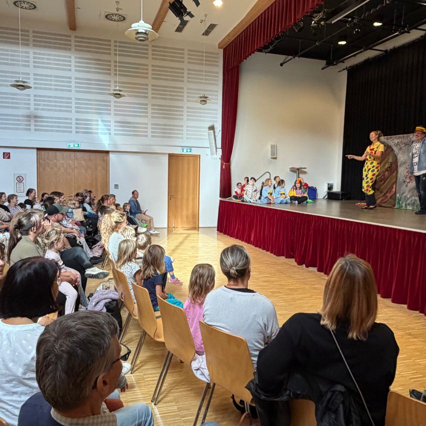 Theateraufführung von Kindern mit Erwachsenen auf der Bühne in einem Saal mit Publikum.
