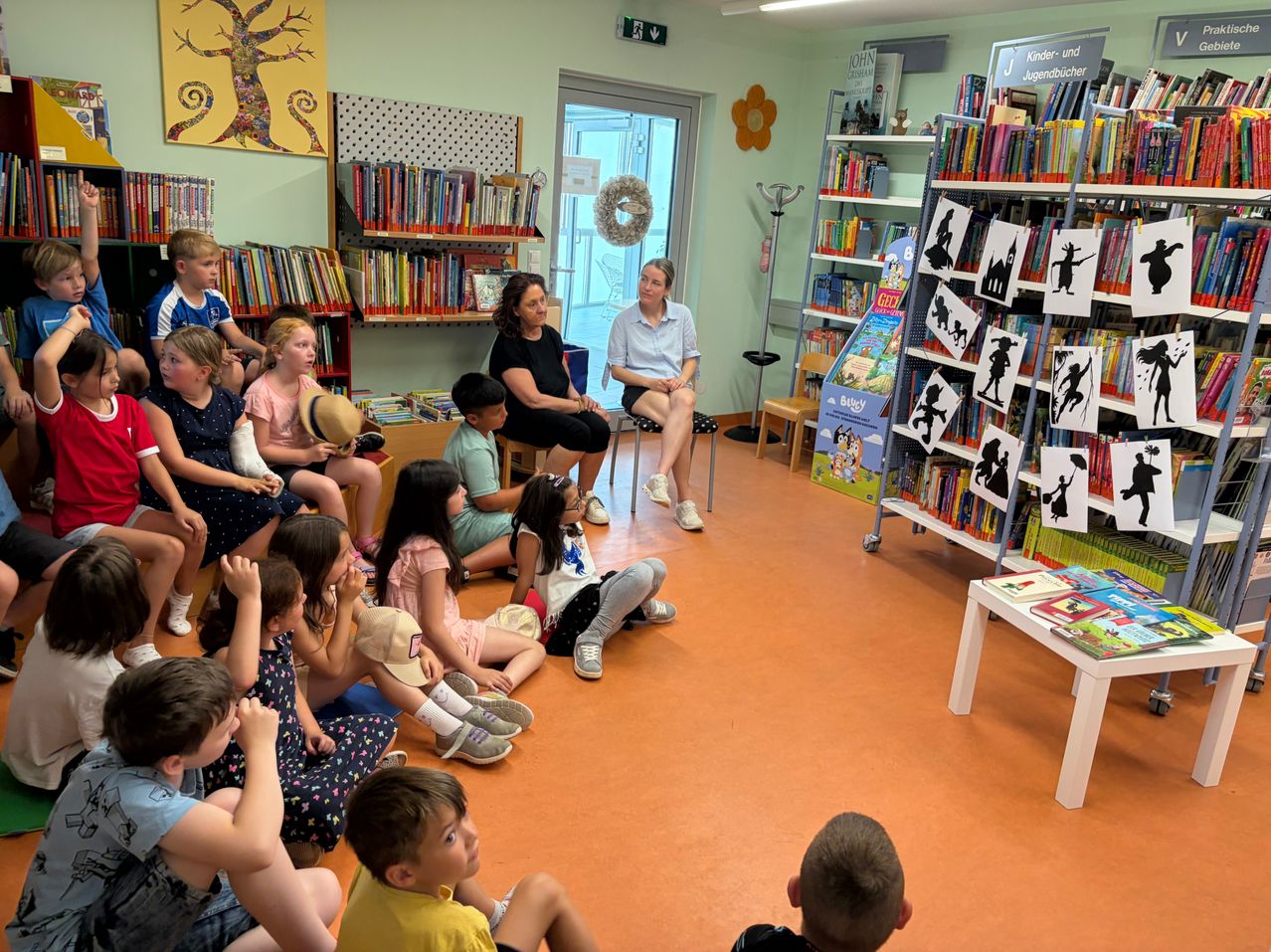 In einer Bibliothek sitzen mehrere Kinder auf dem Boden und hören aufmerksam einer erwachsenen Frau zu, die in einem Stuhl sitzt. Die Bibliothek hat Regale voller Bücher und ein Schild mit Silhouetten steht rechts.