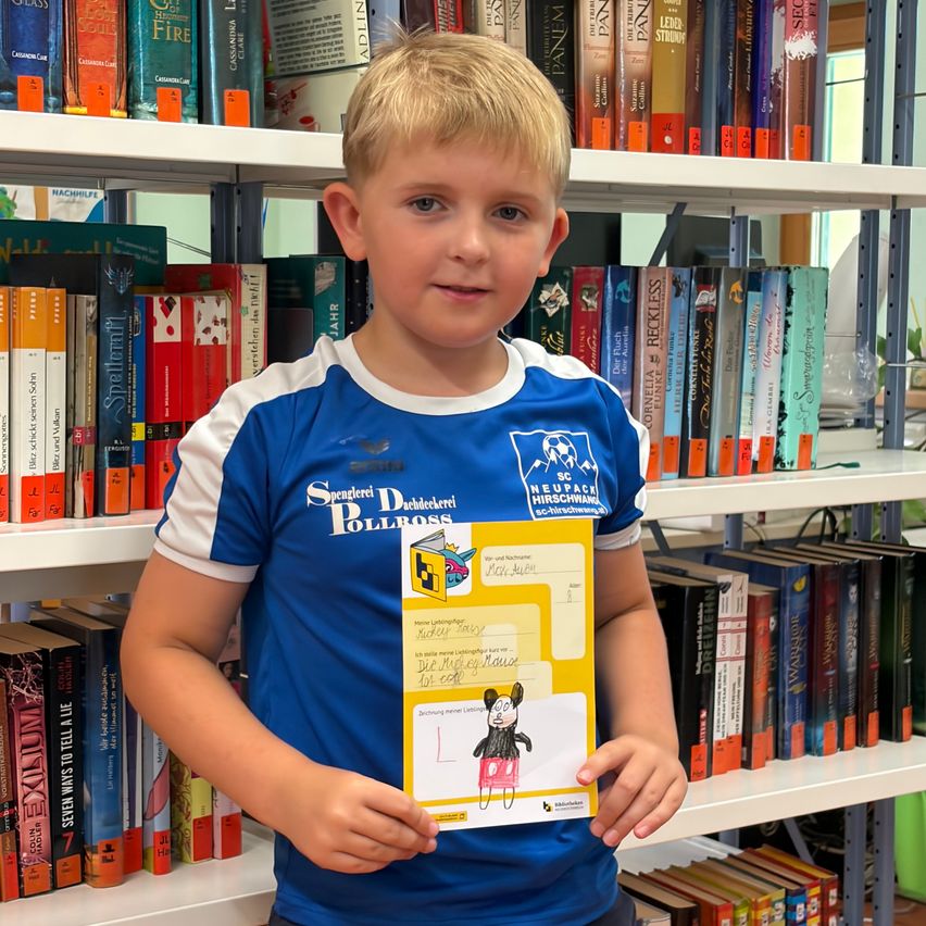 Ein junger Junge steht in einer Bibliothek, trägt ein blau-weißes Fußballtrikot und hält ein Zertifikat. Er lächelt.