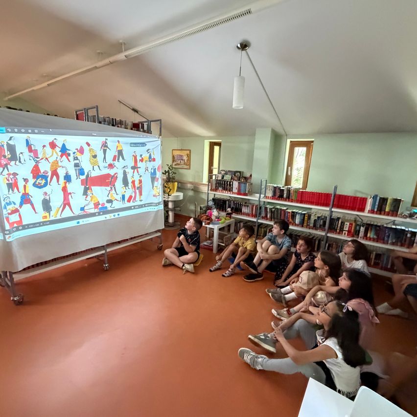Kinder versammeln sich in einer Bibliothek um eine große Projektionsleinwand. Der Raum hat Bücherregale, einen Tisch und eine Deckenlampe.