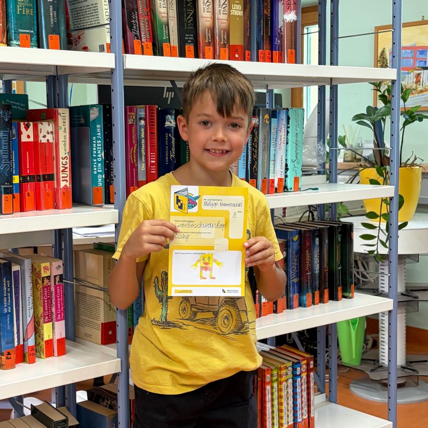 Ein junger Junge steht in einer Bibliothek, lächelt und hält eine Urkunde in den Händen. Hinter ihm sind mit Büchern gefüllte Regale zu sehen.