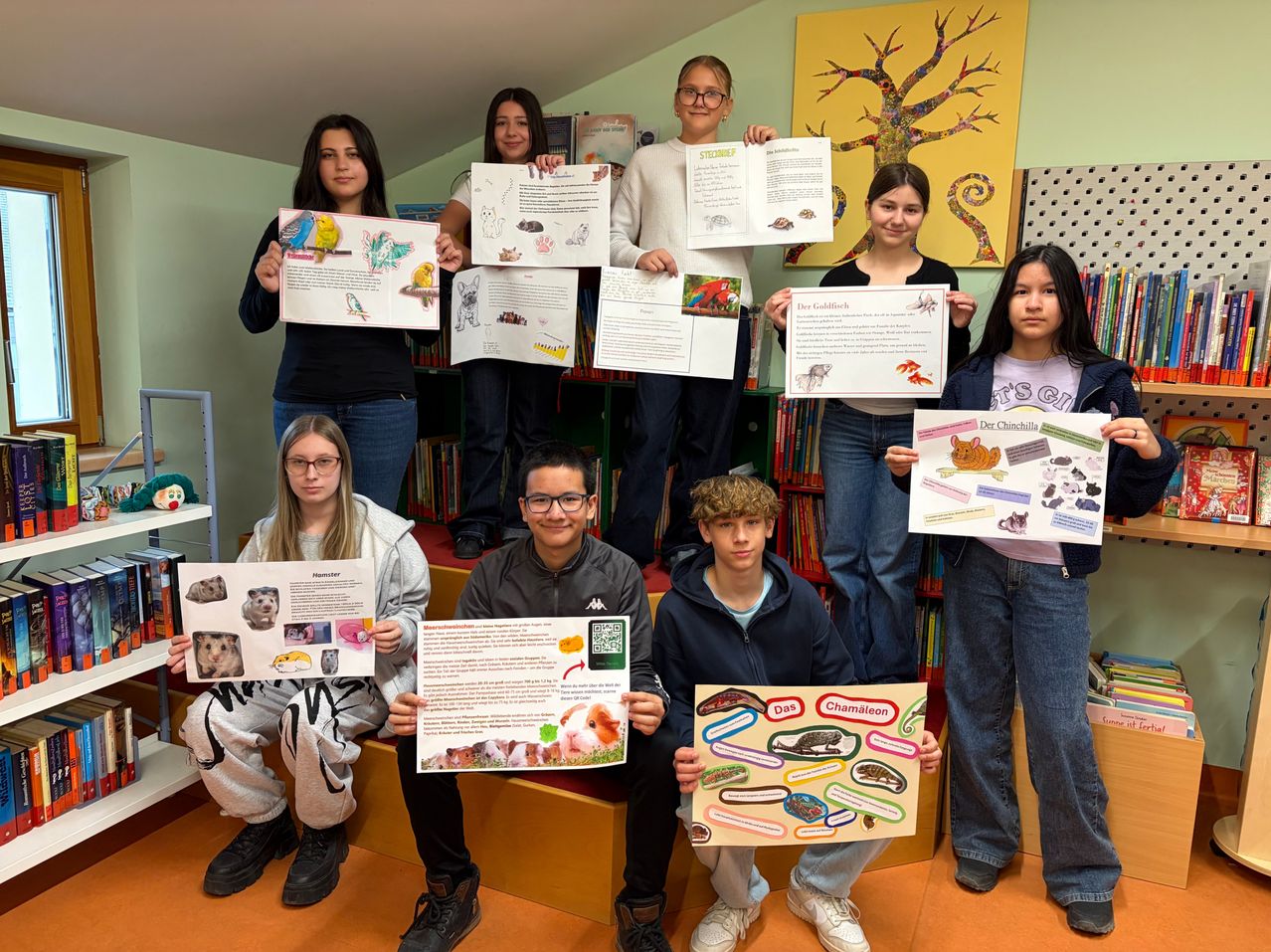 A group of students is holding up posters about animals and pets. The posters feature illustrations of various animals and plants. The students are smiling and seem to be posing for a photo. Behind them is a bookshelf with books and a wall with a tree painting.