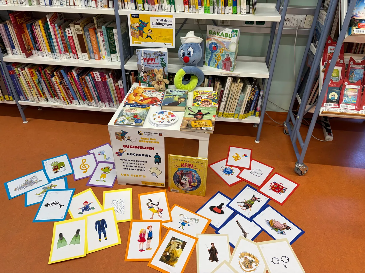 Ein Bibliotheksboden mit einem Tisch voller Bücher, einem Plüschtier und verstreuten Karten. Hinter dem Tisch sind Regale mit Büchern gefüllt.
